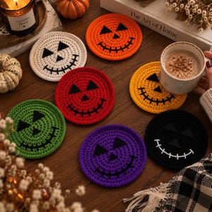 Puede incluir: Un conjunto de siete posavasos circulares de ganchillo con caras de calabaza en naranja, blanco, rojo, amarillo, verde, morado y negro. Cada posavasos tiene ojos triangulares y una boca cosida, con el posavasos negro mostrando costuras blancas.
