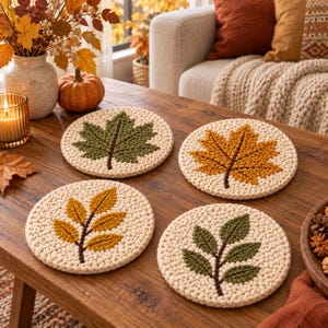 May include: Four round crochet coasters featuring autumn leaf designs. Two coasters display single maple leaves in olive green and golden yellow, while the other two feature branch patterns with smaller leaves in matching yellow and green tones.