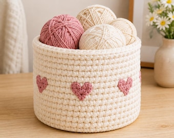 May include: A round, cream-colored crocheted basket featuring a repeating pattern of small, pink heart-shaped accents stitched around the exterior. The basket is filled with four balls of yarn, including one pink ball and three cream-colored balls.