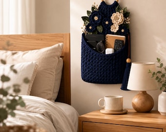 May include: A dark navy blue crocheted wall hanging organizer featuring a wooden ring at the top and decorative crocheted flowers in navy and cream colors. The pocket contains a notebook, a remote control, and other small personal items.