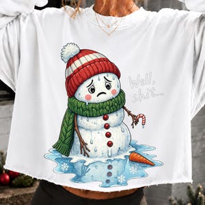Könnte beinhalten: Ein weißes Langarm-Sweatshirt mit einer Grafik eines schmelzenden Schneemanns, der eine rot-weiß gestreifte Mütze und einen grünen Schal trägt. Der Schneemann schaut traurig, hält eine Zuckerstange und der Text "Well, shit..." steht daneben.