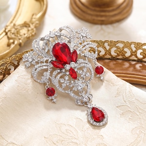 May include: A silver-toned brooch featuring intricate filigree metalwork encrusted with clear rhinestones. The design includes multiple deep red teardrop-shaped crystals and three dangling red crystal charms, creating a symmetrical, ornate appearance.