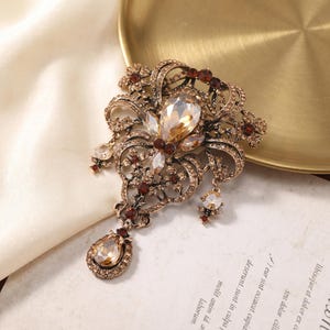 May include: An ornate brooch crafted from antique-toned metal, featuring intricate filigree patterns adorned with numerous small clear crystals, deep red accent stones, and two prominent, teardrop-shaped champagne-colored faceted crystals.