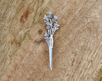 Op de afbeelding: Een zilverkleurige distel haarspeld. De speld heeft een gedetailleerd distelontwerp met bladeren en bloemhoofden. De speld staat op een houten achtergrond.