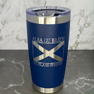 Puede incluir: Un vaso térmico azul marino con borde y tapa plateados. El vaso presenta el texto "ALBA GU BRÀTH" sobre un diseño de bandera escocesa plateada, con "SCOTLAND FOREVER" debajo. El vaso está diseñado para mantener las bebidas frías o calientes.
