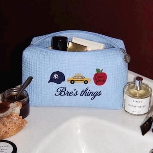 Pode incluir: Uma bolsa azul-clara com textura de waffle e fecho de correr, com ícones bordados de um boné azul-marinho, um táxi amarelo e uma maçã vermelha com o texto New York. Abaixo, o texto Bre's things está bordado numa fonte cursiva azul-escura.