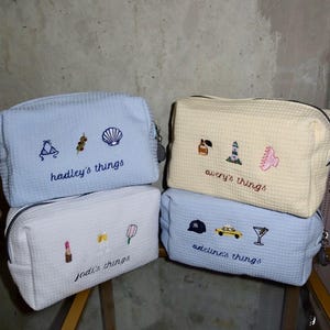 May include: Four rectangular waffle-textured pouches in light blue, white, and cream. Each features embroidered icons and personalised text: 'hadley's things', 'avery's things', 'jodi's things', and 'adeline's things' with motifs like shells, cars, and drinks.
