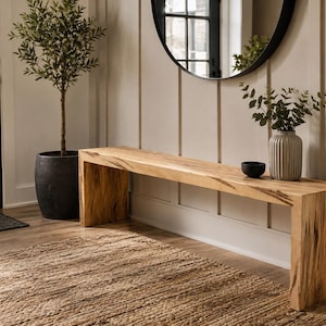 Peut inclure: Un long banc rectangulaire fabriqué en bois clair avec des motifs de grain sombres et prononcés. Le banc présente un design de type cascade où la surface supérieure se prolonge sans interruption en deux supports latéraux verticaux.