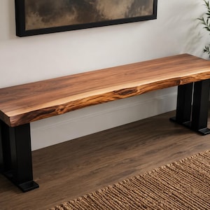 Peut inclure: Un long banc doté d'une assise en bois à bord naturel avec un grain brun riche. L'assise est soutenue par deux pieds en métal noir robustes et rectangulaires placés à chaque extrémité, offrant une base stable pour le plateau en bois.