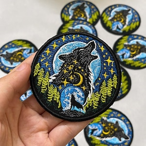 May include: A circular embroidered patch featuring a black wolf howling at a yellow crescent moon against a blue night sky. The design includes yellow stars, a smaller silhouette of a wolf, and green pine trees, all bordered by a thick black edge.
