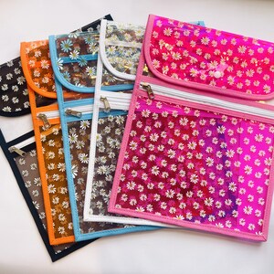 May include: A collection of five rectangular transparent pouches featuring a repeating white daisy pattern. Each pouch has a colored border in black, orange, blue, white, or pink, a top flap with a snap closure, and a horizontal zippered pocket.
