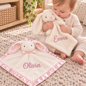 May include: Two cream-coloured plush bunny security blankets with soft fabric. One blanket features the name Olivia embroidered in pink, and the other features the name Emma embroidered in pink. Both have pink satin trim and bunny heads.