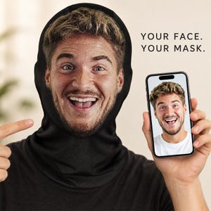 May include: A black fabric face mask featuring a high-resolution printed image of a smiling person with light brown hair. The text 'YOUR FACE. YOUR MASK.' is printed in black on the right. A smartphone displays the same face image.