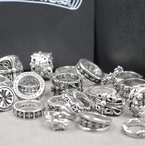 May include: A collection of silver rings featuring intricate embossed cross motifs and engraved text. The rings display various band widths and detailed metalwork, with some pieces showing the Chrome Hearts brand name and 925 sterling silver markings.