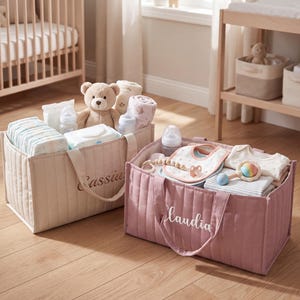 May include: Two rectangular quilted fabric storage baskets with handles. The beige basket features the name Cassius, while the dusty rose basket features the name Claudia. Both contain baby essentials like diapers, bottles, bibs, and plush toys.
