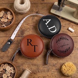 Può includere: Tre metri a nastro retrattili in similpelle con testo personalizzato. Un metro nero presenta le lettere A.D, uno color cuoio mostra le lettere R.T e uno bordeaux riporta il nome Sophia circondato da un disegno a corona di foglie.