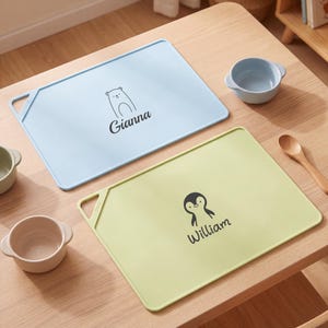 May include: A light blue rectangular silicone placemat with a bear graphic and the name Gianna, and a light green rectangular silicone placemat with a penguin graphic and the name William, each featuring a corner cutout and rounded edges.