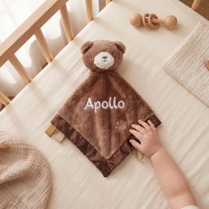 Puede incluir: Una manta de seguridad de felpa marrón con una cabeza de oso de hocico blanco, bordada con el nombre Apollo en hilo blanco. La manta cuadrada tiene un borde de satén marrón oscuro y un pequeño detalle de huella bordado en la tela suave.