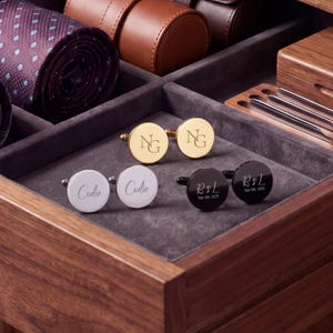 Può includere: Tre paia di gemelli rotondi con finiture oro, argento e nero. La coppia dorata presenta le iniziali NG. La coppia argentata è incisa con il nome Codie. La coppia nera mostra le iniziali R & L con la data Sep 6th, 2025.