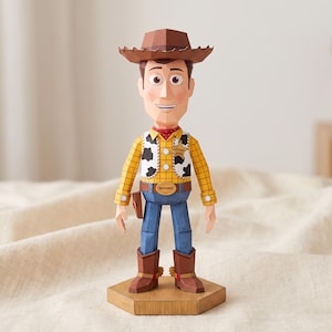 Può includere: Una figura di carta di un personaggio cowboy che indossa un cappello marrone, una camicia a quadri gialla, un gilet bianco con macchie nere, una stella da sceriffo dorata, una cintura blu con una fibbia con la scritta WOODY, pantaloni blu e stivali da cowboy marroni.