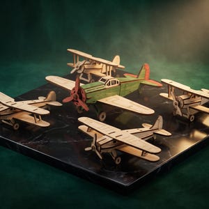 Può includere: Una collezione di cinque modellini di aerei in legno. Un aereo centrale è dipinto di verde con un'elica rossa e dettagli sulla coda, mentre gli altri quattro sono biplani in legno chiaro naturale con eliche e piccole ruote sul carrello.