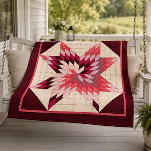 May include: A rectangular quilt featuring a large central star pattern composed of geometric diamond shapes in shades of pink, magenta, and deep burgundy. The star is set against a cream-colored background with a grid pattern and a solid burgundy border.