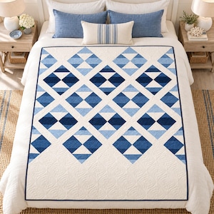 Könnte beinhalten: Eine Steppdecke mit einem geometrischen Rautenmuster in Marineblau, Mittelblau und Hellblau auf cremefarbenem Hintergrund. Die Decke hat einen einfarbigen, cremefarbenen unteren Bereich mit dezenter Steppstruktur und einen dunkelblauen Rand.