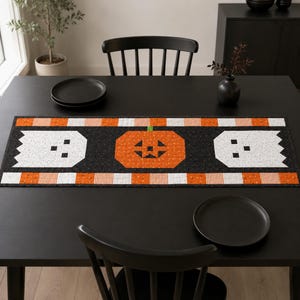 May include: A rectangular fabric table runner featuring a black center panel with a central orange jack-o'-lantern flanked by two white ghosts. The runner is bordered by horizontal stripes of orange, white, and black patterned fabric.