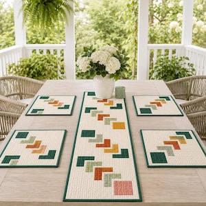 Puede incluir: Un camino de mesa acolchado y cuatro manteles individuales cuadrados a juego con un patrón geométrico de chevrón en tonos verde oscuro, salvia, amarillo mostaza, naranja y rosa claro sobre fondo blanco roto con bordes de tela verde.