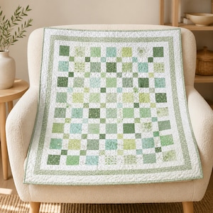 Könnte beinhalten: Eine rechteckige Steppdecke mit einem Patchwork-Design aus verschiedenen grünen und weißen Stoffquadraten. Die Decke hat einen breiten Rand mit einem zarten grünen Blumenmuster und ist durchgehend mit weißen Nähten versehen.