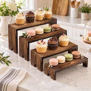 Op de afbeelding: Een set van vier getrapte houten displaystandaards met een donkerbruine gebeitste afwerking. De standaards zijn in een trapsgewijze formatie opgesteld en bevatten diverse cupcakes met glazuur en kleurrijke macarons versierd met kleine witte bloemen.