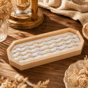 Può includere: Un vassoio espositore per anelli in legno chiaro di forma ottagonale, con un rivestimento interno in velluto bianco. Il vassoio contiene ventiquattro delicati anelli in metallo, ciascuno con una piccola pietra trasparente, disposti in tre file.