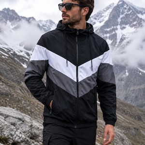 Può includere: Una giacca a vento con cappuccio caratterizzata da un motivo a chevron a blocchi di colore nero, bianco e grigio. La giacca presenta una chiusura frontale con zip, maniche lunghe con polsini elastici e un cappuccio con coulisse, abbinata a pantaloni neri.