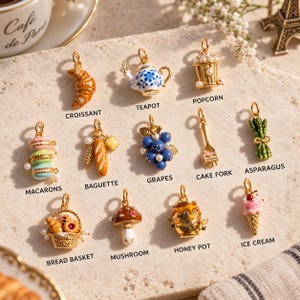 Könnte beinhalten: Eine Sammlung von goldfarbenen Metallanhängern mit Emaille- und Perlenakzenten, darunter Formen wie Croissant, Teekanne, Popcorn, Macarons, Baguette, Weintrauben, Kuchengabel, Spargel, Brotkorb, Pilz, Honigtopf und Eiscreme mit Beschriftungen.
