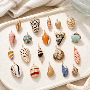 May include: A collection of twenty metal jewellery charms shaped like various seashells. Each charm features colourful enamel detailing in patterns including spots, stripes, and solid hues, with a small gold-toned loop at the top for attachment.
