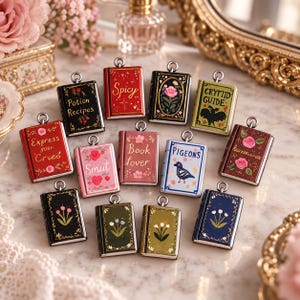 Puede incluir: Una colección de doce dijes de metal rectangulares con forma de libro y anillas plateadas. Cada dije presenta un diseño de portada único, incluyendo títulos como Potion Recipes, Spicy, Book lover, Pigeons y Cryptid Guide, junto con varios motivos florales y estelares.