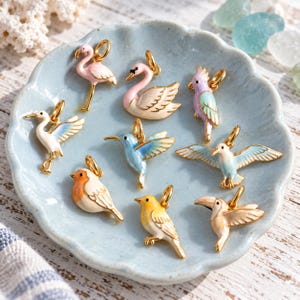 May include: A collection of nine gold-toned metal charms shaped like various birds, including a pink flamingo, a white swan, a purple parrot, a white heron, a blue hummingbird, a seagull, a robin, a yellow songbird, and a toucan.
