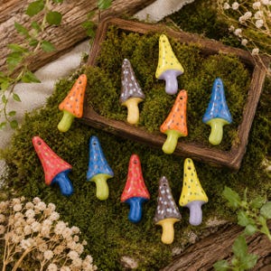 May include: A collection of ten small ceramic mushroom pendants in various colors including yellow, orange, red, blue, and brown, each featuring a spotted cap and a solid-colored stem with a small hole at the top for hanging or stringing.