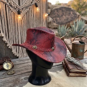 May include: A red and black snakeskin-patterned cowboy hat featuring a braided hatband adorned with two circular metallic studs. The hat is displayed on a black mannequin head, showcasing the textured material and the structured brim design.
