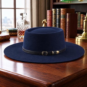 May include: A navy blue flat-brimmed hat with a stiff, cylindrical crown. The hat features a thin black textured band encircling the base of the crown, secured with a small, rectangular brass-toned metal buckle and a matching metal tip.