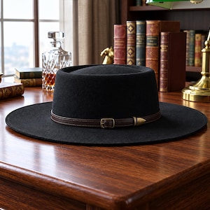 May include: A black flat-brimmed hat with a flat crown, featuring a dark brown leather band around the base. The band includes a small, rectangular brass-toned metal buckle and a pointed metal tip, providing a structured and clean appearance.