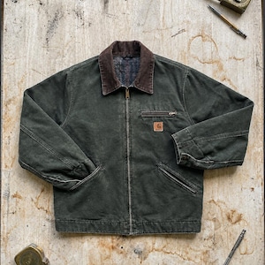 Può includere: Una giacca in tela verde scuro con colletto in velluto a coste marrone, chiusura frontale con zip integrale e una piccola toppa quadrata con logo Carhartt sul petto. La giacca presenta una tasca sul petto con zip, due tasche inferiori e maniche lunghe.