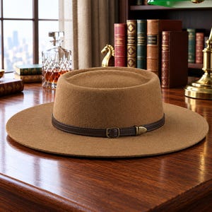 Può includere: Un cappello in feltro color marrone chiaro con corona piatta e tesa larga. Il cappello presenta una fascia in pelle marrone scuro attorno alla base della corona, fissata con una piccola fibbia in metallo e una punta metallica affusolata.
