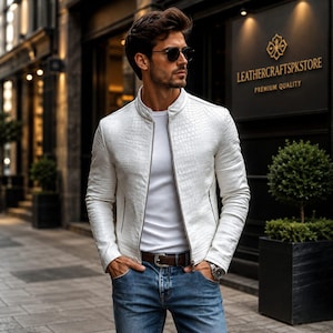 May include: A white jacket with a textured crocodile skin pattern, featuring a stand-up collar, a full-length front zip, and two side pockets. The jacket is worn over a plain white t-shirt, paired with blue denim jeans and a brown leather belt.