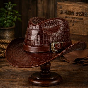 Op de afbeelding: Een donkerbruine cowboyhoed met een textuur van krokodillenleer. De hoed is voorzien van een gladde bruine leren band rond de kroon, vastgezet met een versierde, antiek ogende koperen gesp. De rand van de hoed is aan de zijkanten omhoog gebogen.