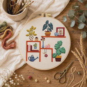 Könnte beinhalten: Ein runder Stickrahmen mit einem Stoffeinsatz, der ein handgesticktes Motiv von orangefarbenen Regalen zeigt, auf denen verschiedene Topfpflanzen, Bücher, eine blaue Gießkanne, ein kleines Haus, Kerzen, ein Bilderrahmen und ein hängender Vogel stehen.