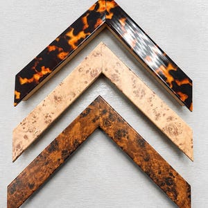 May include: Three V-shaped picture frame corner samples arranged vertically. The top sample features a glossy tortoiseshell pattern in brown and orange. The middle sample has a light beige burl wood grain. The bottom sample shows a dark brown burl wood.