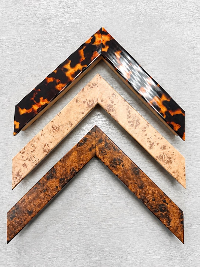 May include: Three V-shaped picture frame corner samples arranged vertically. The top sample features a glossy tortoiseshell pattern in brown and orange. The middle sample has a light beige burl wood grain. The bottom sample shows a dark brown burl wood.