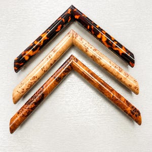 Puede incluir: Tres muestras de esquinas de marcos de fotos en forma de V dispuestas verticalmente. El marco superior tiene un patrón de carey brillante en marrón oscuro y naranja. El marco central es de madera veteada beige claro. El marco inferior es de madera veteada marrón rojiza.