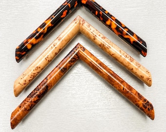 May include: Three V-shaped picture frame corner samples arranged vertically. The top frame has a glossy dark brown and orange tortoiseshell pattern. The middle frame is a light tan wood burl. The bottom frame is a rich reddish-brown wood burl.